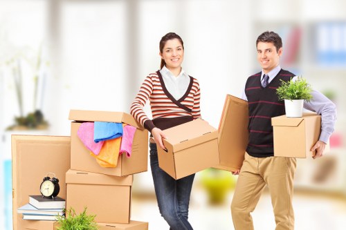 A man and woman standing inside a bright, modern residential space with large windows, surrounded by several cardboard boxes of various sizes, some sealed and others open, revealing household items. The woman, dressed in a red and white striped sweater and jeans, holds a medium-sized box, smiling at the camera. The man, wearing a dark sweater with stripes over a shirt and beige trousers, holds two smaller boxes and a potted plant with green foliage, also smiling. In the background, there are additional boxes stacked nearby, a small black clock, and a leafy green plant, all set on a light wooden floor. The scene conveys an ongoing house removal or relocation process, with a neutral and orderly atmosphere typical of professional removals in the Canary Wharf or London Docklands area. The overall lighting is bright and natural, emphasizing the clean, contemporary interior environment suitable for house removals services.