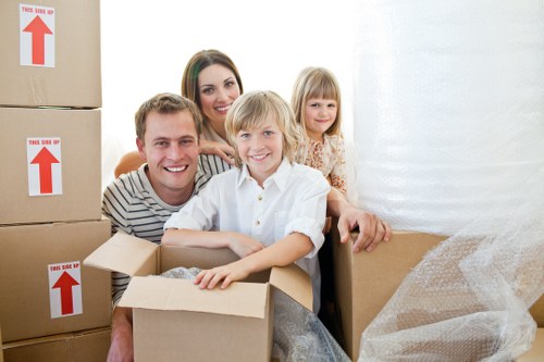 A family of four, including a father, mother, and two young children, are inside a residential space surrounded by packed cardboard boxes and bubble wrap, indicating they are in the process of moving house. The father, wearing a striped shirt, is leaning forward with a smile, while the mother, with long dark hair, stands behind him, also smiling. The boy, with blond hair, is sitting with his arms resting on an open cardboard box filled with packing material, and the girl, with light-colored hair, stands slightly behind them next to a large cardboard box covered with bubble wrap. Several packed cartons, some labeled with red arrows, are stacked nearby, suggesting professional house removals. The environment features a white wall background and natural lighting, with a residential hallway or room setting typical of a home preparing for a move, supporting the context of local removals services in Canary Wharf or nearby.