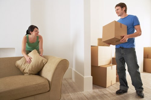 A young woman with dark hair tied back, wearing a green sleeveless top, is seated on a beige sofa inside a residential property, holding a large beige cushion on her lap and smiling as she looks towards a man. The man, dressed in a blue T-shirt and dark jeans, is standing nearby holding a cardboard moving box with both hands, preparing to place it among several other stacked cardboard boxes in the background. The interior space features light-colored walls and wooden flooring, with natural light illuminating the scene. The setting appears to be a living room in a house or apartment, indicative of a typical house removal or relocation process, with visible boxes and furniture suggesting an ongoing move. This image reflects a professional approach to house removals services that Canary Wharf Man with Van provides, emphasizing careful handling of household items during a move in or around the Canary Wharf area, postcode E14, in East London.