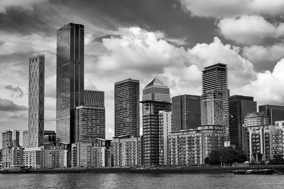 A high-angle view of a cityscape at sunset showing a cluster of tall modern skyscrapers with reflective glass facades in Canary Wharf, London. The image captures the urban environment with multiple of