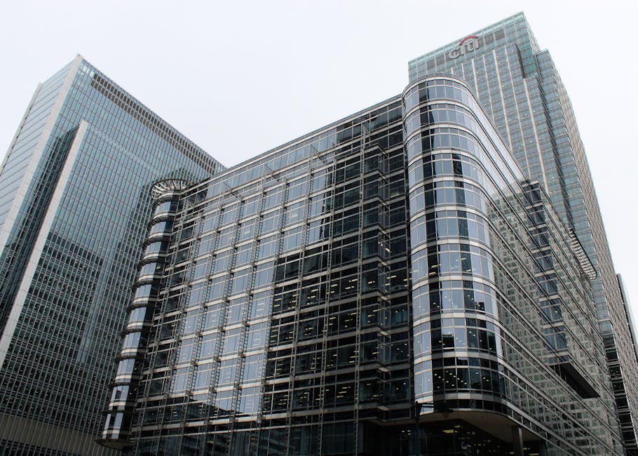 A cityscape at dusk showing tall modern office buildings with glass facades reflecting the evening sky, situated along a canal with calm water. The buildings vary in height and architectural style, wi