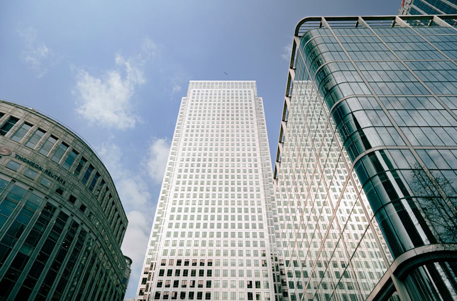 Inside a modern office building with large floor-to-ceiling glass windows revealing a cityscape of tall office towers and blue skies. The entrance area features a glass revolving door with metal frami