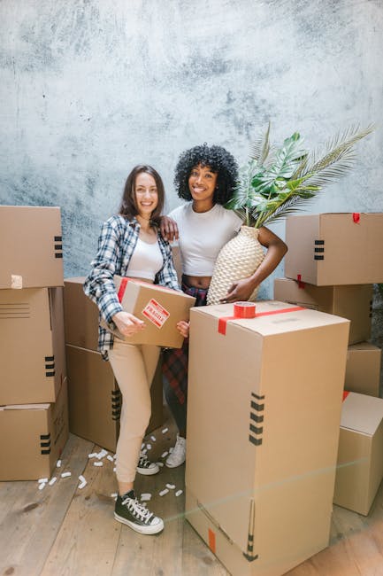 A person holding an open cardboard box with a red and white 'Fragile' handle with care sticker, standing among several stacked boxes in an indoor setting with blue patterned wallpaper. The individual�
